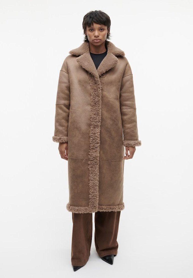 Bruine nepleren shearlingjas met een grote kraag, voorknoopsluiting en getextureerde afwerking. Model draagt brede bruine broeken en puntige zwarte hakken.