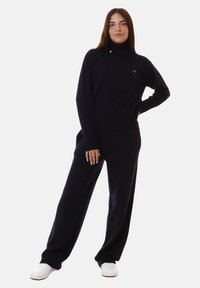 Maglione a collo alto blu navy con polsini e orlo a costine, abbinato a pantaloni a gamba larga in coordinato. Entrambi realizzati in tessuto morbido. Footwear bianco visibile.
