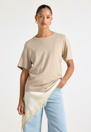Kvinde med mørkt hår iført beige kortærmet top, lyseblå jeans og hvidt halstørklæde med blonder bundet om livet, stående mod hvid baggrund.
