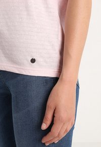 Camisa de manga corta texturizada color rosa claro con un acento redondo oscuro en el dobladillo, combinada con jeans de mezclilla azules, que se muestra parcialmente.