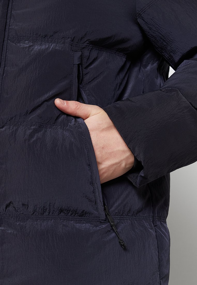 Marine kleur pufferjack met een gestructureerde afwerking, met een hand die in een zijzak reikt en een koord aan de onderkant voor aanpassing.