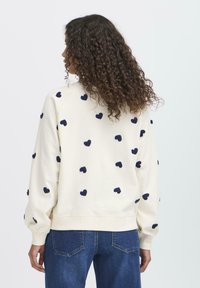 Felpa color crema con ricamo a forma di cuore blu navy, polsini e orlo a coste, caratterizzata da una vestibilità ampia e una texture morbida. Indossata con jeans.