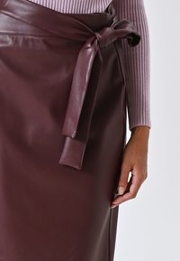 Gonna avvolgente in pelle bordeaux con dettaglio a cravatta, caratterizzata da una texture liscia e una silhouette aderente. Sono visibili le costine di una maglietta viola chiaro.