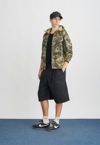 Kamikraāsu mēteļi ar melniem akcentiem, melnas īsas bikses, melna cepure un kedas. Mēteļiem ir rāvējslēdzēja priekšpuse un sānu kabatas.