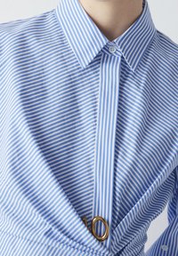 Camicia a righe blu chiaro e bianco con colletto bottonato e un accento dorato. Presenta un design avvolgente e un tessuto testurizzato.