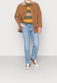 Man som bär en brun jacka över en randig grön, orange och beige skjorta, ljusblå jeans och mångfärgade sneakers med vita snören, står med en hand i fickan.