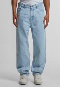 Jeans din denim de culoare albastră deschisă, cu o croială relaxată, având fermoar, cinci buzunare și o ușoară decolorare. Purtați cu adidași albi.