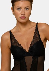 Bodysuit en dentelle noire avec des motifs floraux, bonnets souples, panneaux en mesh transparent, bretelles ajustables et accents en métal doré.