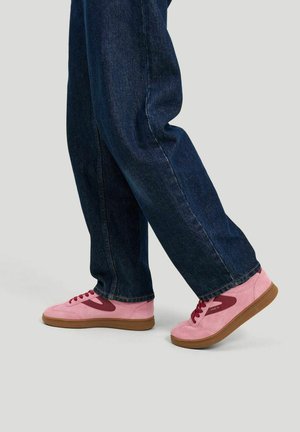 Henkilö, jolla on tummansiniset farkut ja vaaleanpunaiset lenkkarit ruskeilla pohjilla, seisoo yksinkertaista vaaleanharmaata taustaa vasten.