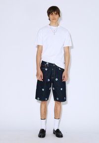 Camiseta de algodón blanca, pantalones cortos de mezclilla oscura con bordados florales, mocasines negros y calcetines blancos, sobre un fondo liso.