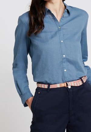 Mujer con una camisa azul de manga larga con botones, metida dentro de un pantalón oscuro con un cinturón estampado en rosa y beige, manos en los bolsillos.