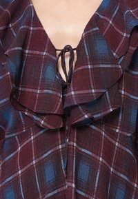 Μπλούζα με καρό σε μπορντώ και μπλε χρώμα, με φούστα λαιμό και κλείσιμο με κορδόνι. Διαθέτει υφασμάτινη υφή με μαλακό, στρωτό σχέδιο.