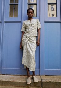 Lichtgekleurde, gestreepte tweedelige outfit met korte mouwen en een strak rokje. Model draagt beige instapschoenen en poseert tegen een blauwe achtergrond.