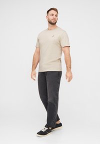 Beige T-Shirt mit kurzen Ärmeln und Rundhalsausschnitt, kombiniert mit dunkelgrauen Jeans und schwarzen Sneakers mit weißen Schnürsenkeln und einer hellen Sohle.