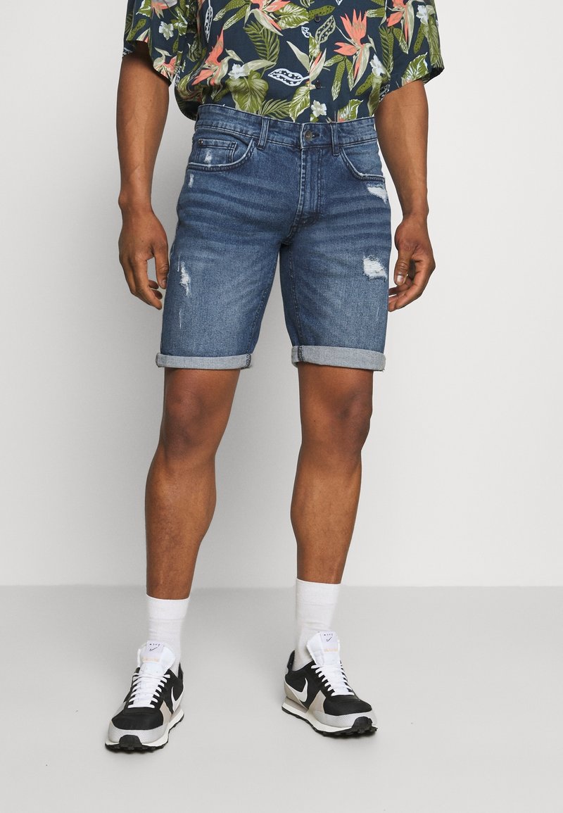 Pantalones cortos de mezclilla en azul oscuro con dobladillos enrollados, con detalles desgastados y bolsillos frontales. Combinados con una camisa de estampado tropical y zapatillas.