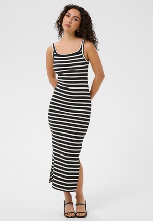 Femme aux cheveux bouclés portant une robe midi sans manches à rayures horizontales noires et blanches et des sandales noires à lanières, debout les mains derrière le dos.