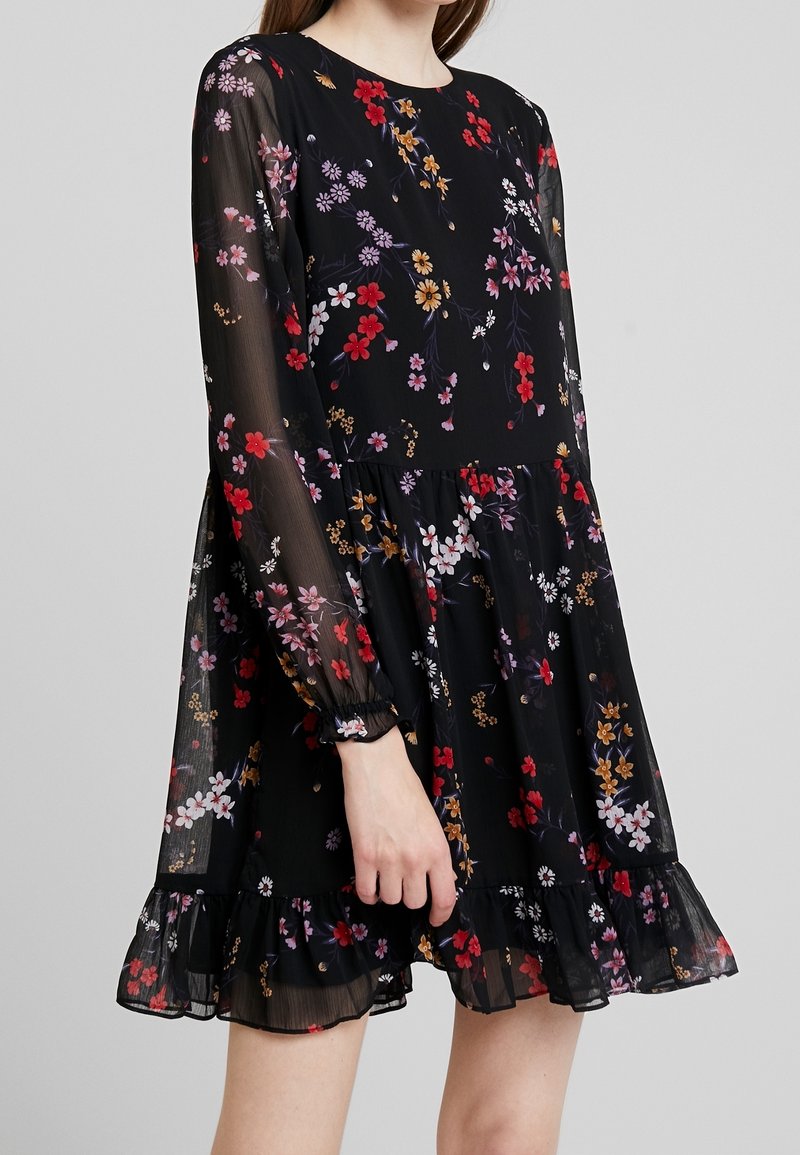 Schwarzes Blumenkleid mit langen, durchsichtigen Ärmeln, das bunte Blumenmuster in Rot, Lila und Gelb zeigt. Der Volantsaum verleiht dem Kleid eine zusätzliche Textur.