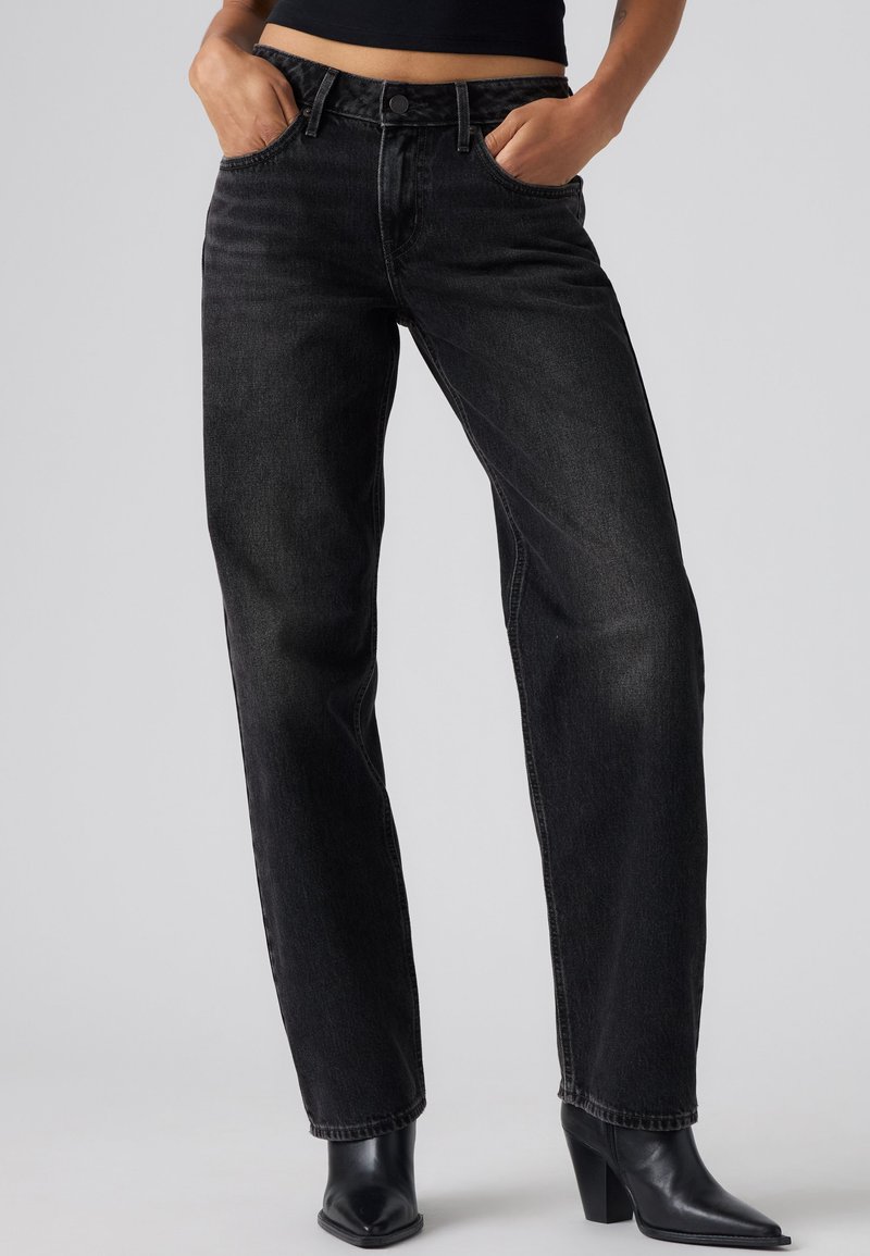 Personne portant un jean noir taille haute à jambes droites, mains dans les poches et bottes à talons noires à bouts pointus sur un fond uni.