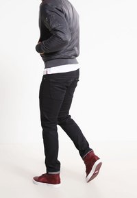 Veste bomber noire, t-shirt blanc à col rond, jean noir et baskets montantes rouges. Tissu lisse, coupe ajustée et couches contrastantes.