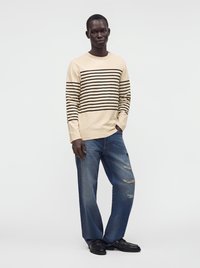 Homme portant une chemise beige à manches longues avec des rayures horizontales noires et un jean bleu déchiré et ample, debout avec une main dans la poche.