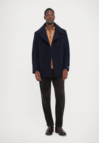 Peacoat de lã em azul-marinho com design de duplo peito e grandes botões pretos, combinado com um suéter bege com fecho, calças de cotim escuras e botas castanhas.