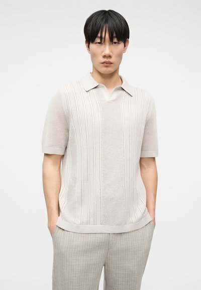 Νεαρός άνδρας με μαύρα μαλλιά που φοράει ένα ανοιχτό μπεζ πλεκτό πόλο με κοντά μανίκια και ανοιχτό μπεζ παντελόνι με ρίγες, στέκεται με τα χέρια στις τσέπες.