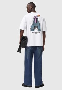 Hvid oversized t-shirt med grafisk design på ryggen, der viser en farverig næve og en rose, ledsaget af løse blå jeans.