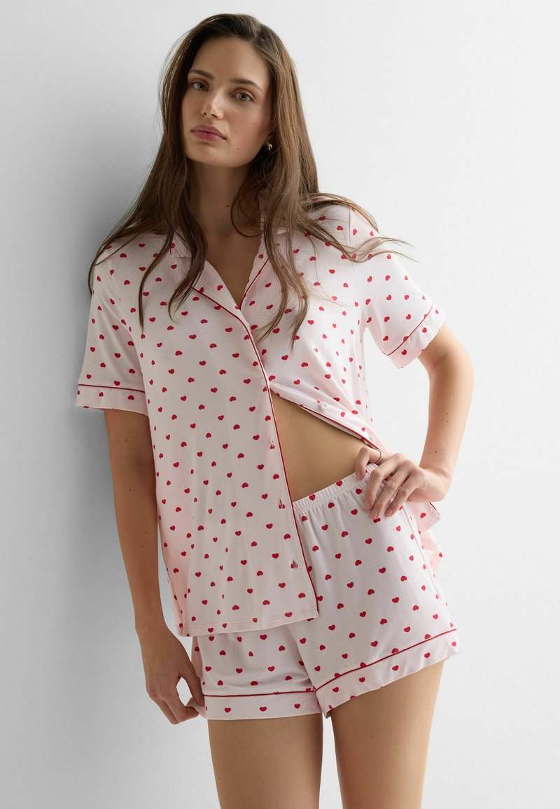 Frau trägt hellrosafarbenes, kurzärmeliges Button-up-Hemd und passende Shorts mit kleinen roten Herzmustern, steht vor weißem Hintergrund.