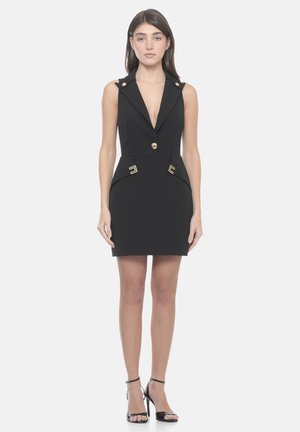 Donna in piedi con un mini abito nero senza maniche con bottoni dorati e sandali neri con tacco alto, su uno sfondo semplice.