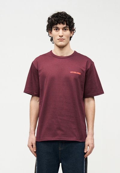 Νέος άνδρας με σγουρά μαλλιά, φορώντας ένα μαύρο T-shirt με κοντά μανίκια και μικρό φωτεινό πορτοκαλί κείμενο στο στήθος, καθώς και σκουρόχρωμο τζιν, στέκεται μπροστά σε λευκό φόντο.