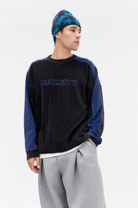 Sweatshirt de velludo corduroy preto com detalhes azuis, apresentando o texto "JETS FRANS". Acompanhado por calças de fato plissadas cinza claro e um gorro com padrão de estrelas azuis.