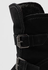 Primer plano de una bota de ante negra con dos hebillas plateadas y un suave forro interior visible en la parte superior.