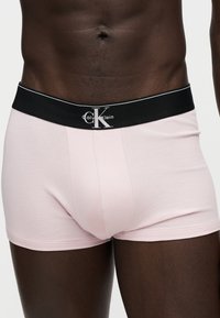 Άνδρας κορμός που φορά ανοιχτό ροζ boxer Calvin Klein με μαύρη μέση και λευκό λογότυπο.