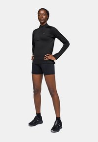 Haut noir à manches longues zippé avec une coupe ajustée, associé à un short de sport noir et des chaussures noires. Texture lisse, design minimal, sans motifs visibles.