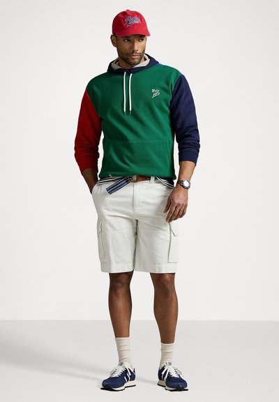 Hoodie coloré en blocs de vert, rouge et marine avec cordon de serrage, associé à un short beige clair, des chaussettes blanches et des baskets bleues. Casquette rouge avec logo.