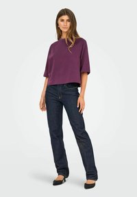 Femme portant un haut violet ample, un jeans bleu foncé, des escarpins noirs à bouts pointus, ainsi qu'un bracelet et des bagues dorées, se tenant contre un fond clair.