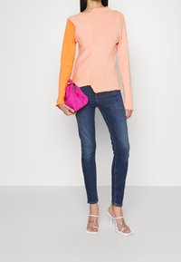 Färgglad stickad tröja i persika och orange, kombinerad med mörkblå denimjeans. Håller en ljusrosa texturerad handväska; bär klara blockklackar.