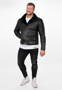 Zwarte leren motorjack met een bontkraag, geboriginaliseerde mouwen, een ceintuur in de taille, ritssluitingen en gedragen met een wit t-shirt en versleten zwarte spijkerbroek.