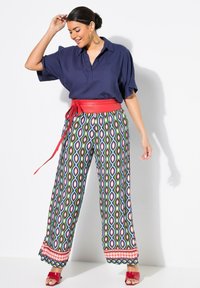 Blauwe blouse met korte mouwen, rode riem, brede broek met geometrisch patroon in groen, blauw en rood, en rode hoge sandaaltjes.