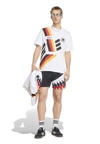 Νέος άνδρας που φοράει λευκό μπλουζάκι Adidas και μαύρο σορτς με κόκκινα, κίτρινα και μαύρα γεωμετρικά σχέδια, κρατώντας ταιριαστό μπουφάν και φοράει γυαλιά ηλίου.