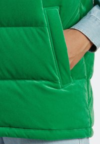 Grön vadderad väst med en mjuk, sammetliknande textur, utrustad med en sidficka och en kontrasterande blå denimskjorta som sticker fram under.