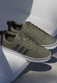 Par de zapatillas Adidas verde oliva con rayas negras y suelas blancas colocadas sobre una superficie geométrica blanca con luz suave.