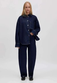 Donkere denim-shirt en wijde jeans, beide met een losse pasvorm. Het shirt heeft een opgestikte zak en knoopdetails. G gedragen met zwarte schoenen.