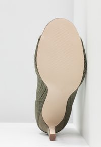 Chaussure tricotée verte avec une surface texturée et un bout pointu. Présente une semelle lisse de couleur claire avec des nervures pour l'adhérence.