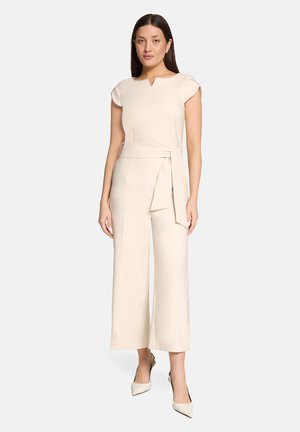 Vrouw met stijlvol donker haar die een crèmekleurige jumpsuit met korte mouwen, een taillekoord en wijd uitlopende cropped broek draagt, gecombineerd met crèmekleurige hakken.