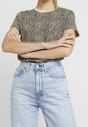 Person trägt ein kurzärmeliges Shirt mit Leopardenmuster und kleinem rotem Levi's-Logo, hellblaue hoch taillierte Jeans, die Arme verschränkt.
