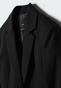 Musta bleiseri, jossa on lovettu kaulus, rakenteelliset hartiat ja sileä kangas. Sisäpuolella on huomaamaton vuori ja brändilabeli. Minimalistinen muotoilu.