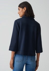 Veste cropped marine avec un large col, des manches 3/4 et des coutures structurées, assortie à un jean bleu ajusté. Matière lisse avec une texture minimale.