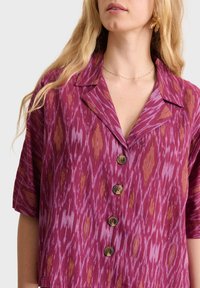 Camisa de manga corta con cuello en magenta profundo con un patrón ikat que presenta tonos amarillos y morados. Los botones son marrones con un diseño floral.