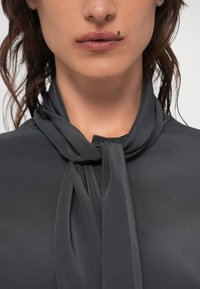 Dunkelgraue Bluse mit einer weichen, glänzenden Textur, die eine geknotete Schleife am Kragen und ein glattes, minimalistisches Design aufweist.